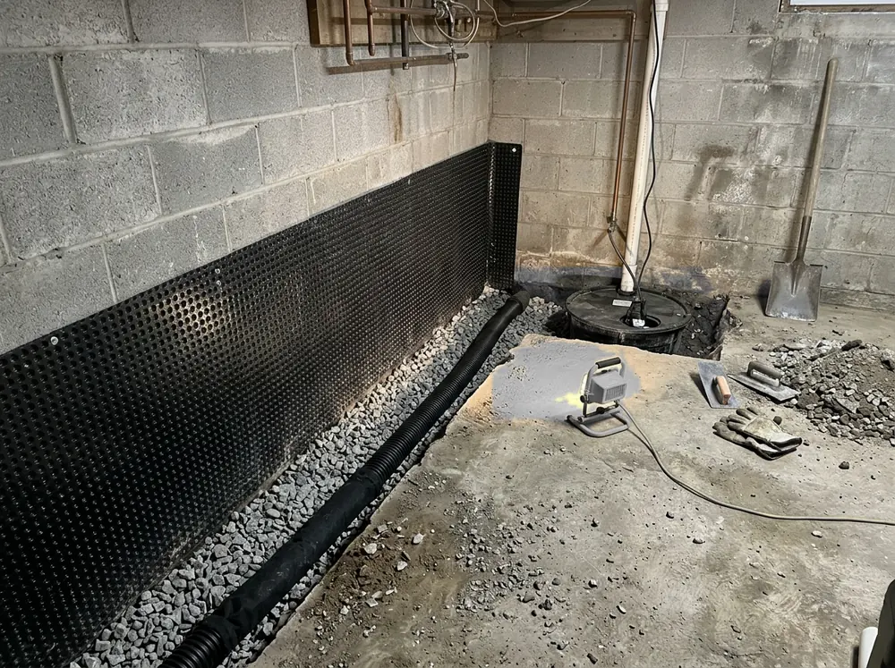 Basement floor opened in a narrow trench along a block wall, gravel and black perforated pipe in the trench, a plastic drainage membrane on the lower wall and a sump basin in the corner