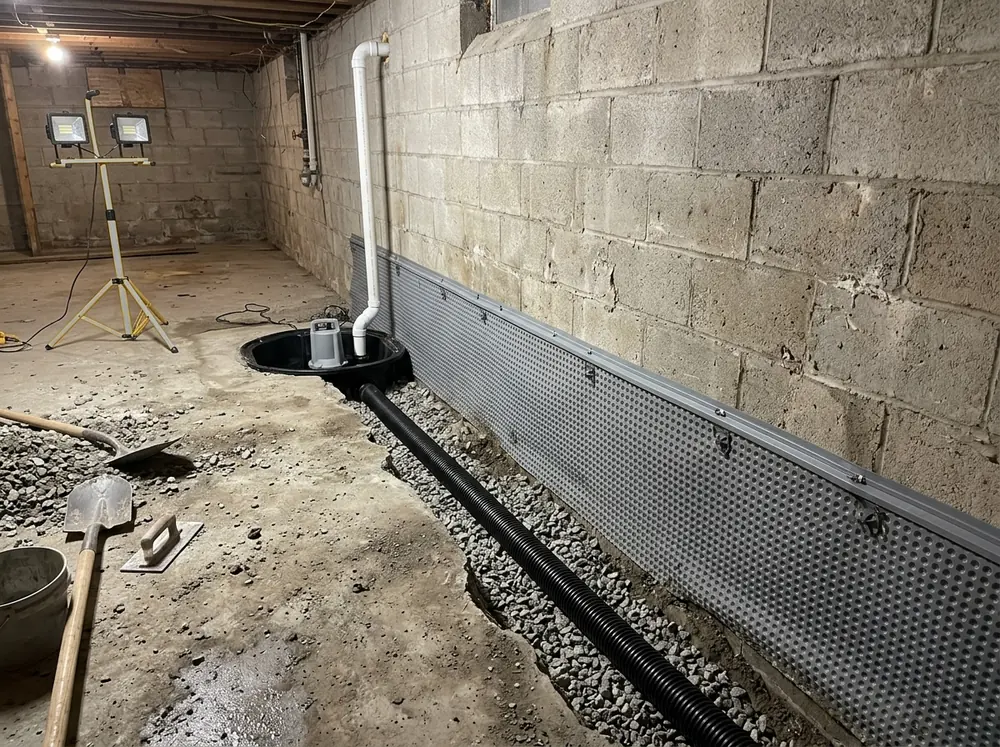 Basement floor trench along a block wall with gravel and black perforated drain pipe, a dimpled drainage membrane on the wall and a new sump basin with a grey pump in the corner