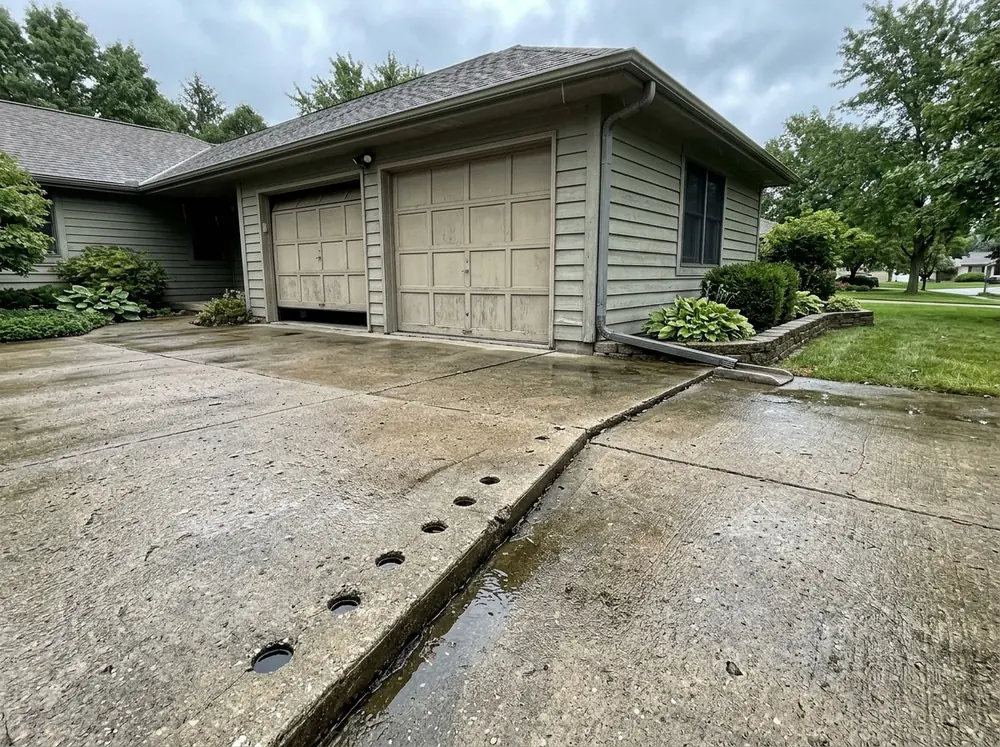 Concrete driveway leading to a two-car garage with one panel sunk about two inches below the next, small round drilled holes in the low panel and water pooled along the joint